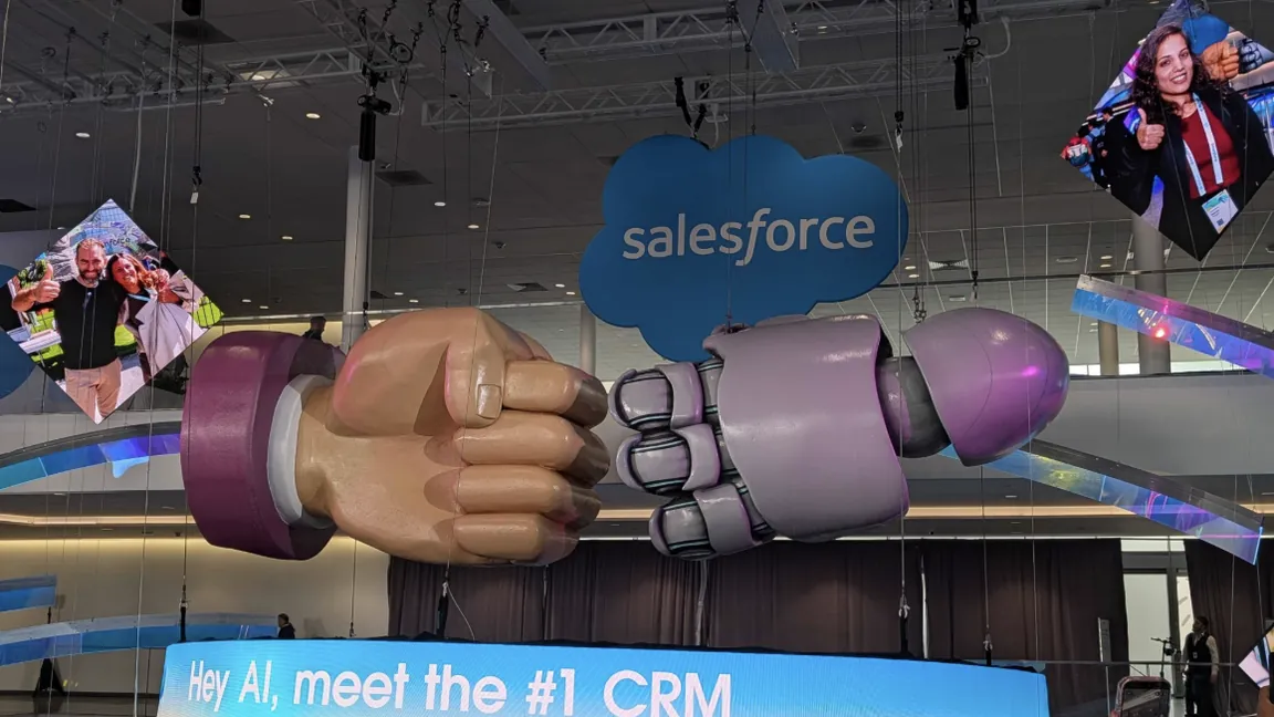 A giant human fist and robot fist bumping under the Salesforce logo above a screen reading "Hey AI, meet the #1 CRM" at Dreamforce 2026